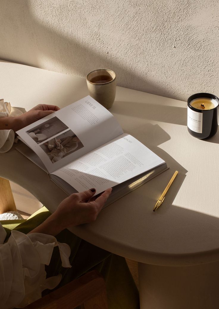 Hands turning the pages of an open magazine beside a candle and cup on a sunlit table